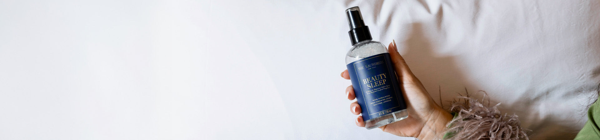 The Laundress Beauty Sleep Fabric Spray for sleepwear and bedding is held over a bed sheet.