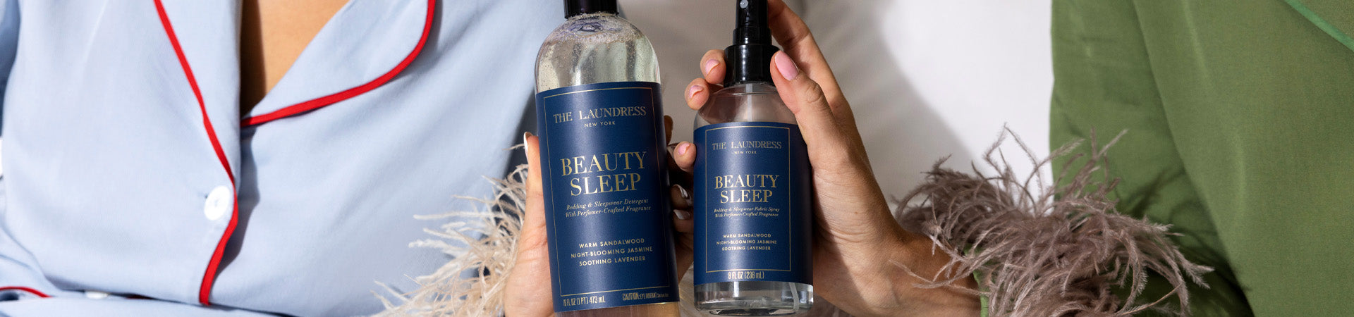 Two people in pajamas with feathered sleeves hold The Laundress Beauty Sleep Bedding & Sleepwear Detergent & Fabric Spray.