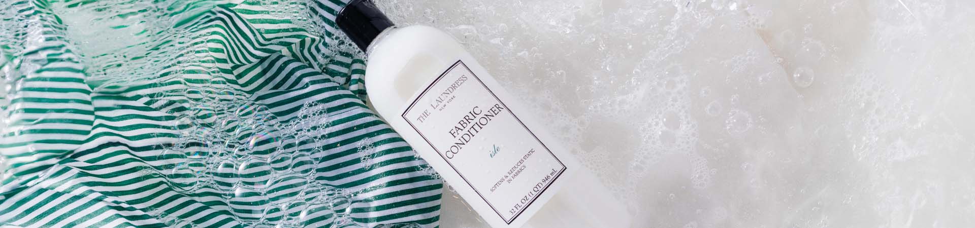 The Laundress Fabric Conditioner in the scent Isle held over sudsy water with a striped fabric in it.