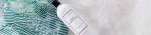 The Laundress Fabric Conditioner in the scent Isle held over sudsy water with a striped fabric in it. 