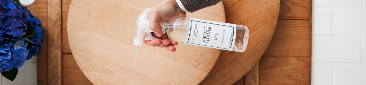The Laundress Surface Cleaner is sprayed onto a wooden surface in a well-lit kitchen with blue flowers nearby.