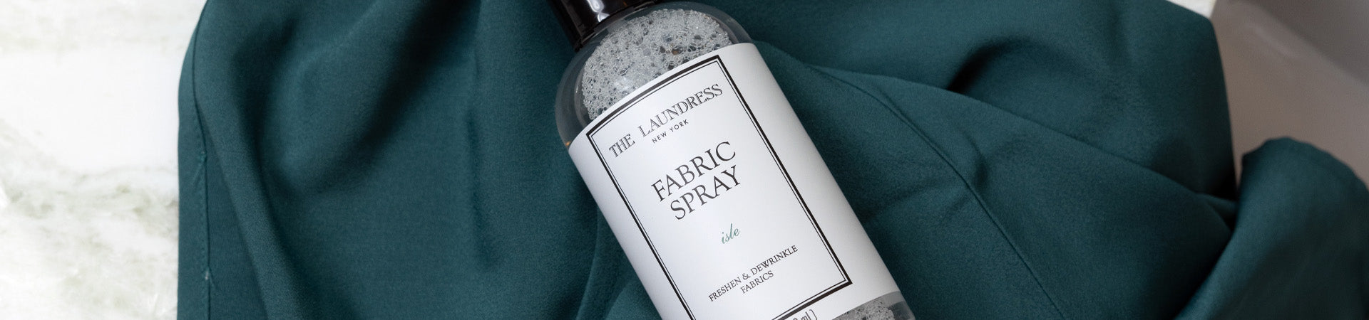 The Laundress Fabric Spray in the coastal-inspired scent Isle lays on a teal fabric on a marble surface.