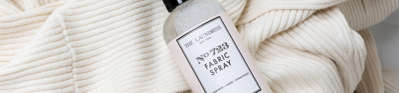 The Laundress No. 723 Fabric Spray, with scents of Damask rose, geranium, and amber, lays on a white knit garment.