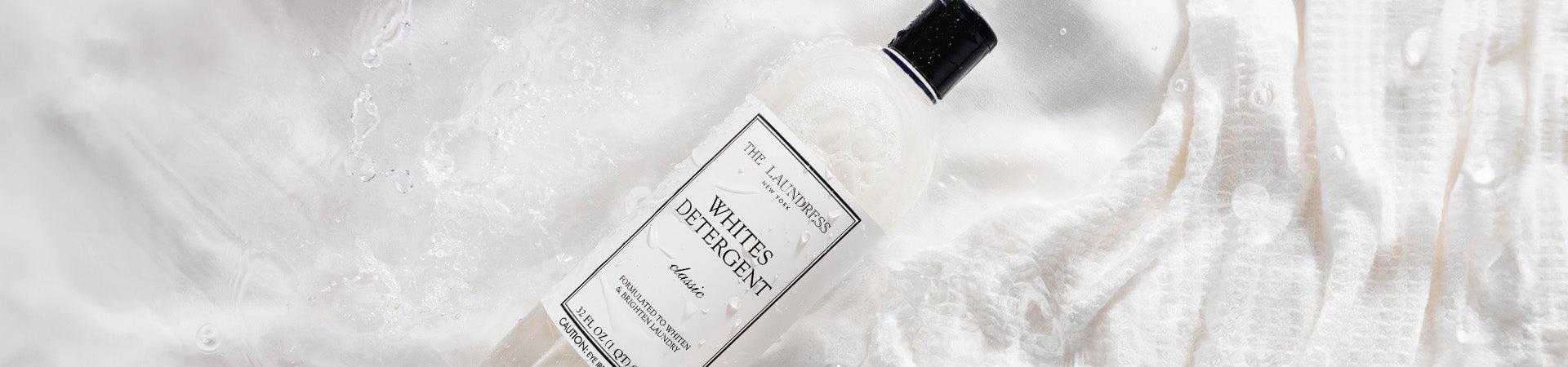 The Laundress Whites Detergent formulated to whiten & brighten laundry laying in water with white fabric.
