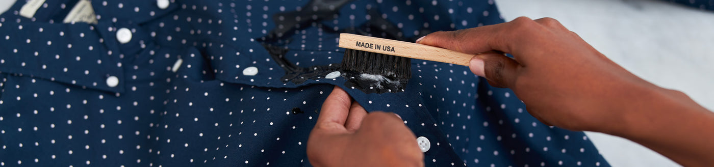 A close-up of The Laundress Stain Brush with soft, densely packed bristles being used to fight a stain on a blue collared shirt. 